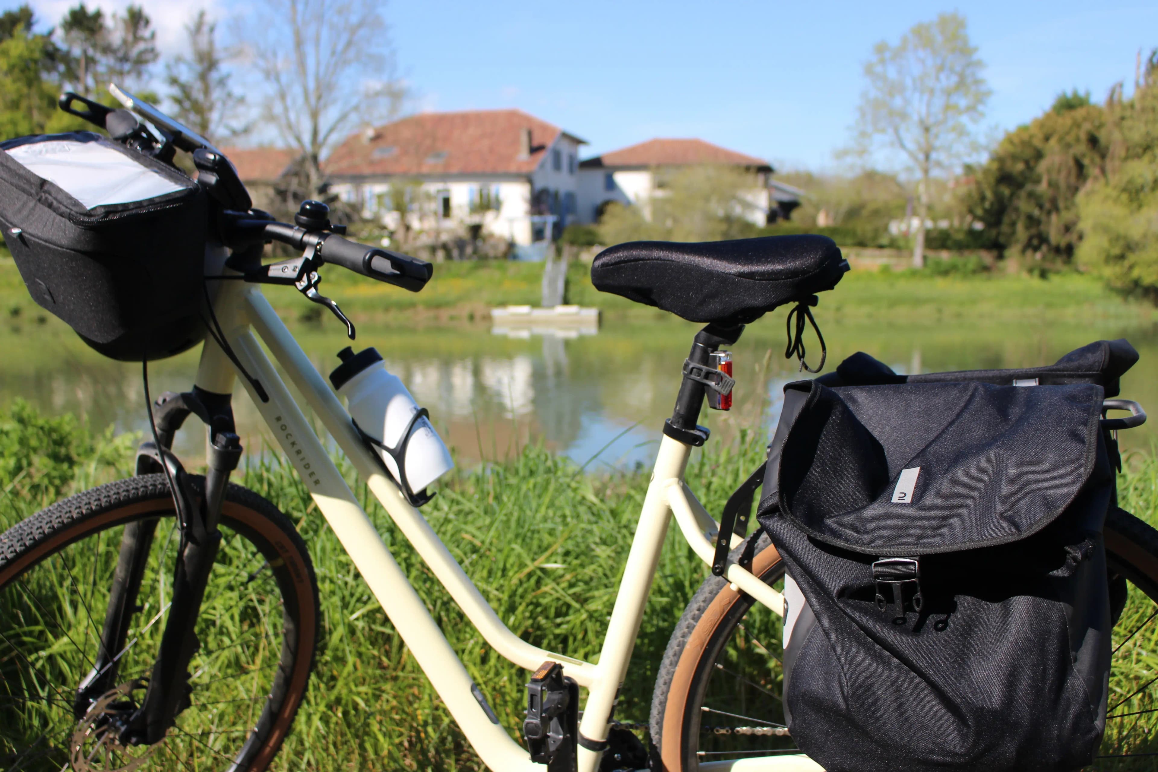 Cream white hiking bike with black panniers and water bottle sitting in front of a pond with houses and trees in the background.
Travel bike rental with The Family Bike agency.