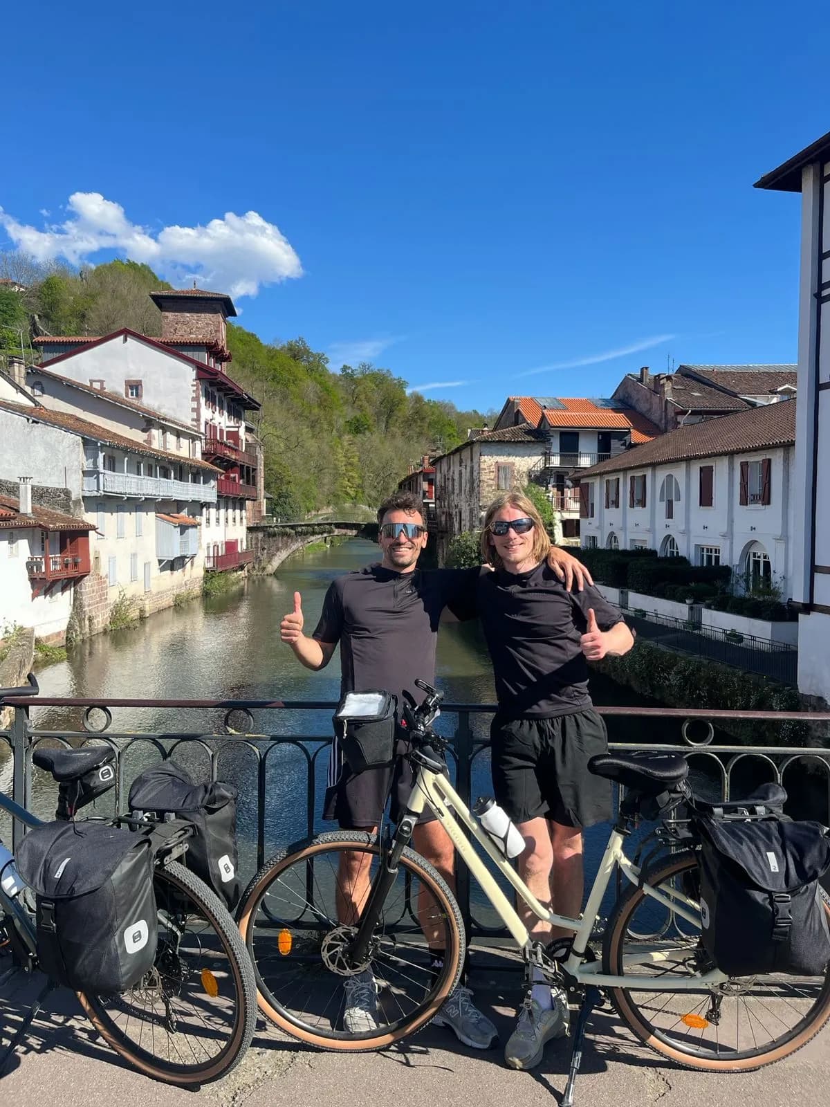 Two men in cycling gear pose with their bikes on a bridge in Saint Jean Pied de Port above a river, with traditional houses and a blue sky in the background.